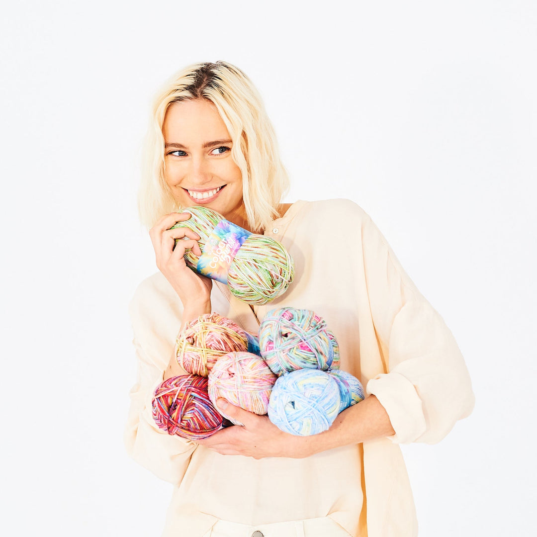A smiling blonde woman in a cream-colored top holds a pile of colorful variegated yarn balls against a plain white background.