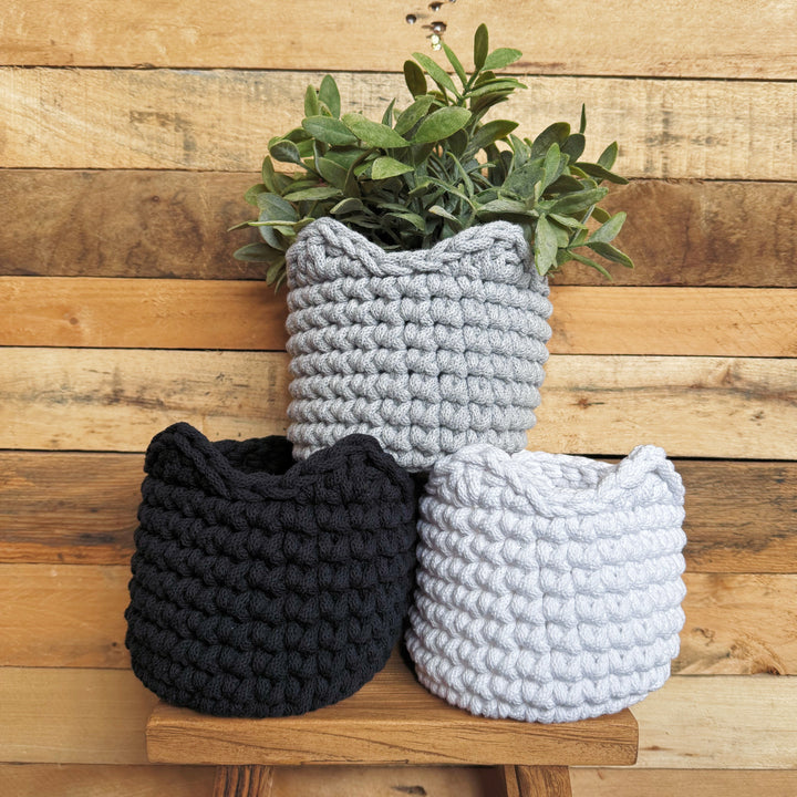 Three knitted planters in black, gray, and white on a wooden surface with a wooden wall background.