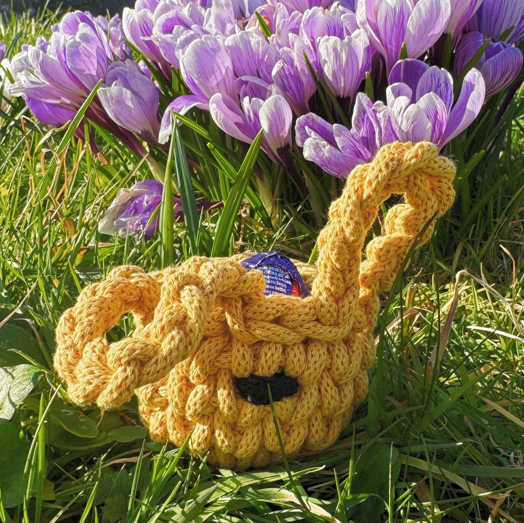 A handmade yellow crocheted Easter bunny basket with long, floppy ears, sitting in grass in front of blooming purple crocuses. The basket holds a foil-wrapped chocolate egg, with bright spring sunlight illuminating the scene.