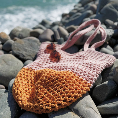 orange and pink crochet string beach bag with sunglasses resting on top, lying on a bed of pebbles