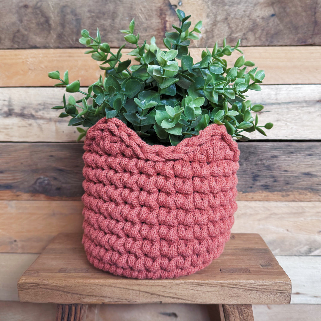 A chunky crocheted planter in a terracotta colour, filled with green eucalyptus foliage, displayed on a rustic wooden stool against a wooden plank wall.