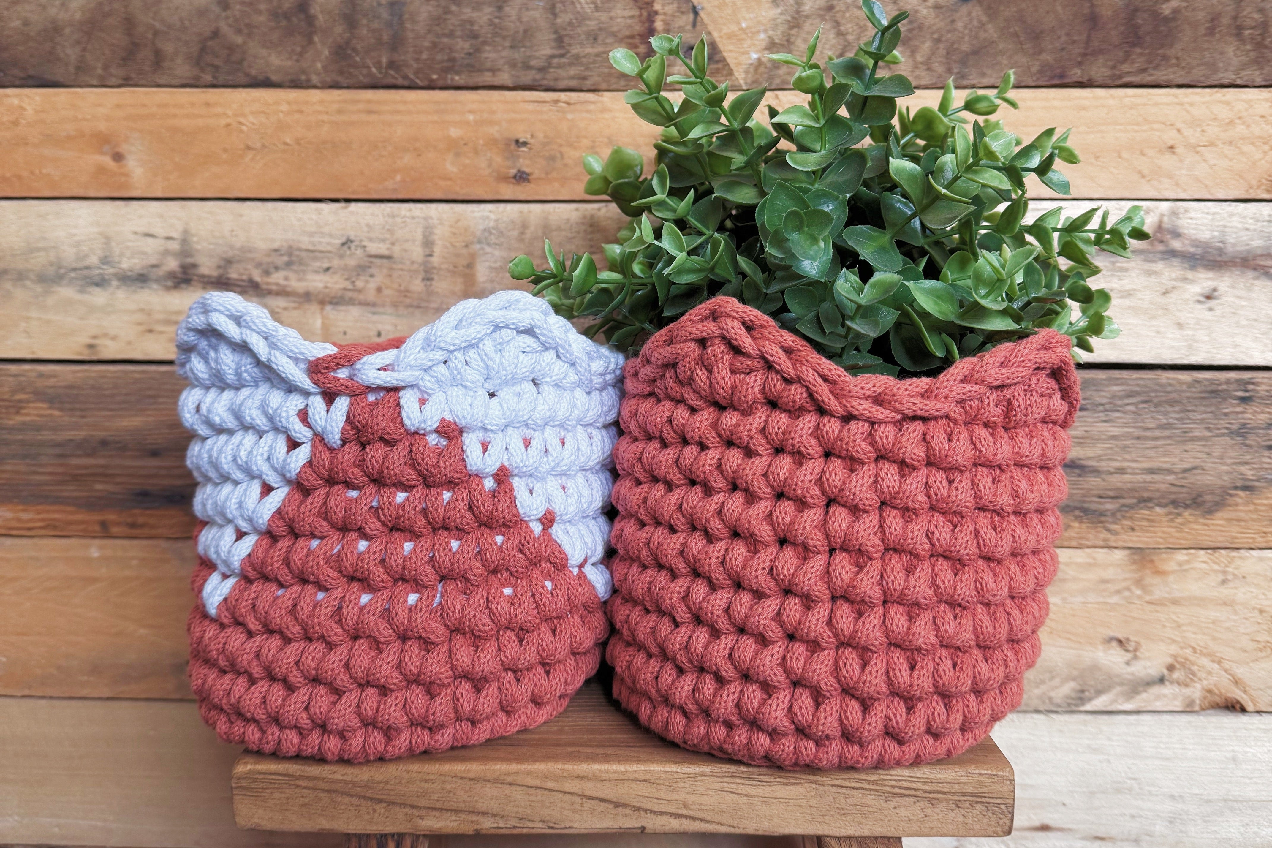 Two crocheted planters with a plant on a wooden stool against a wooden wall.