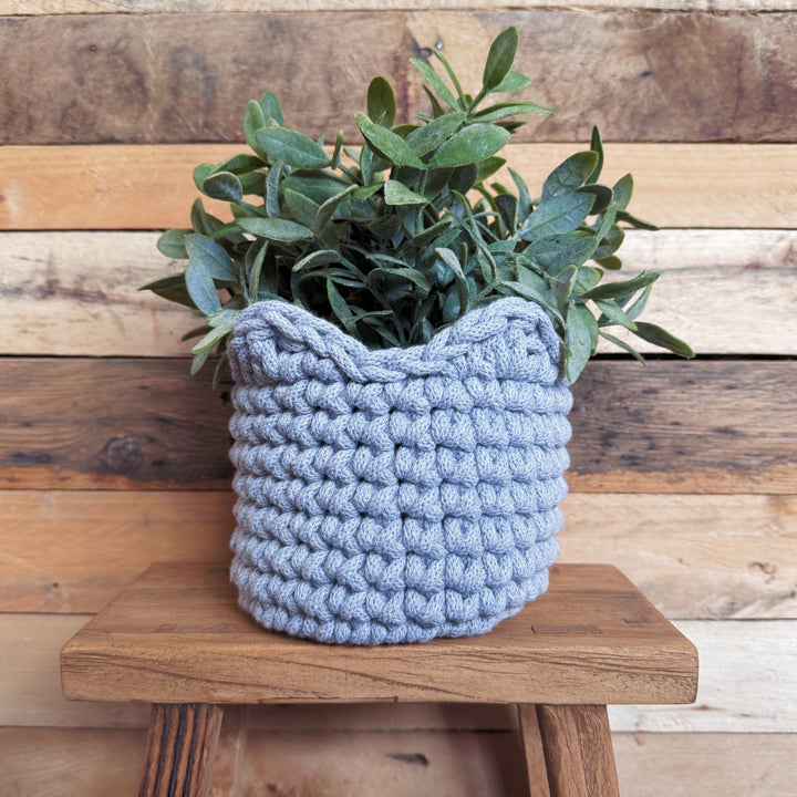 A light grey crocheted planter basket holding green foliage, displayed on a rustic wooden stool against a wood plank wall.