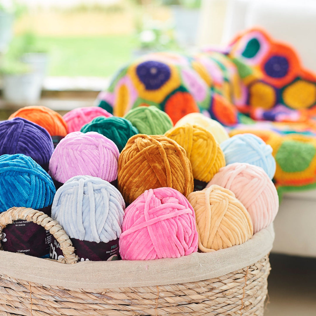 A woven basket filled with colorful balls of yarn in shades of blue, pink, purple, green, yellow, orange, and beige, with a crocheted blanket in the background on a sofa.