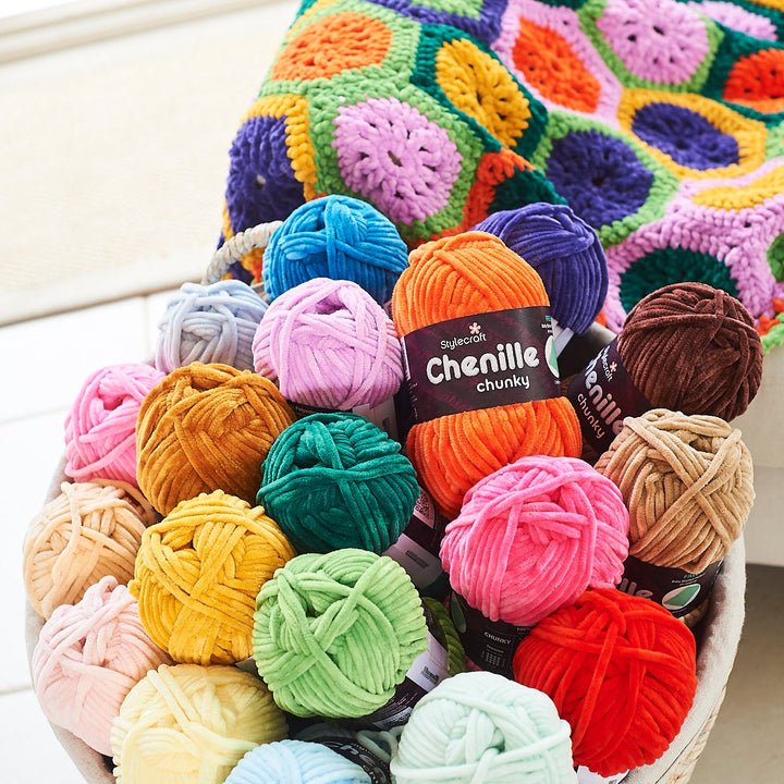 A woven basket filled with colorful balls of chenille yarn, with a crocheted granny-square blanket draped over the basket. A label on one orange ball reads &