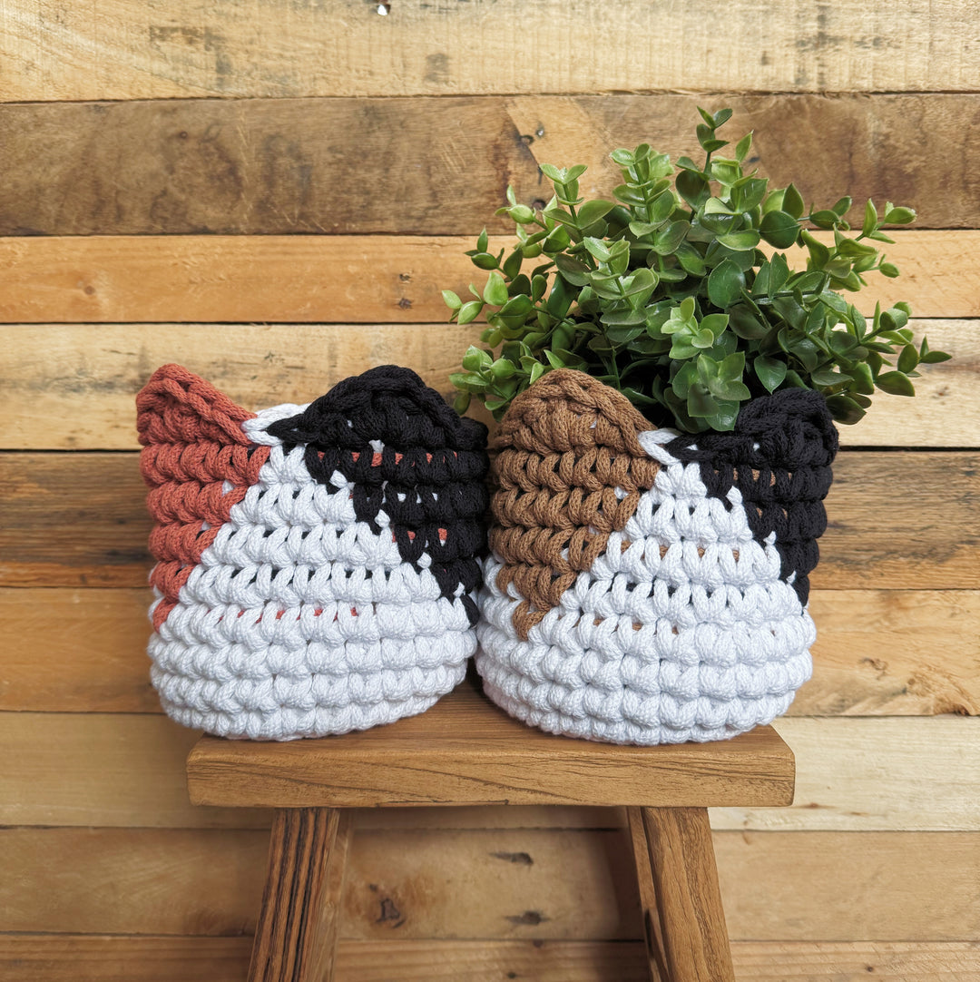 Two crocheted planter baskets shaped like cat heads, one in white, black, and terracotta, and the other in white, black, and brown, holding green plants and displayed on a wooden stool against a rustic wood plank wall.