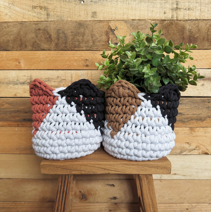 Two crocheted planter baskets shaped like cat heads, one in white, black, and terracotta, and the other in white, black, and brown, holding green plants and displayed on a wooden stool against a rustic wood plank wall.