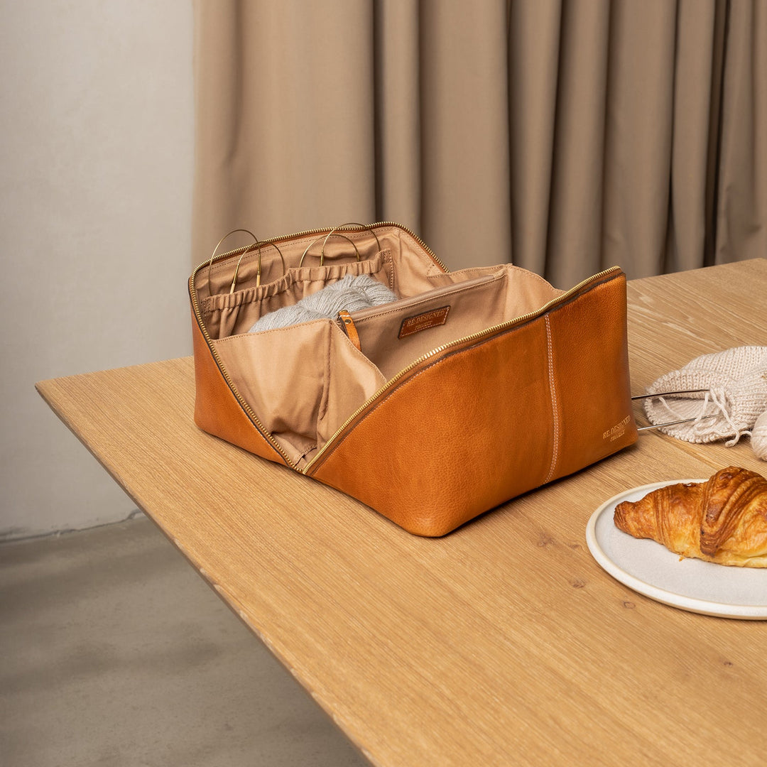 Brown leather bag on a wooden table with a plate of croissants and white fabric in the background.