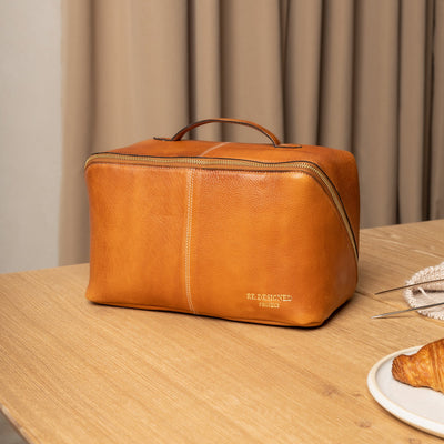 Brown leather toiletry bag on a wooden table with a neutral curtain background