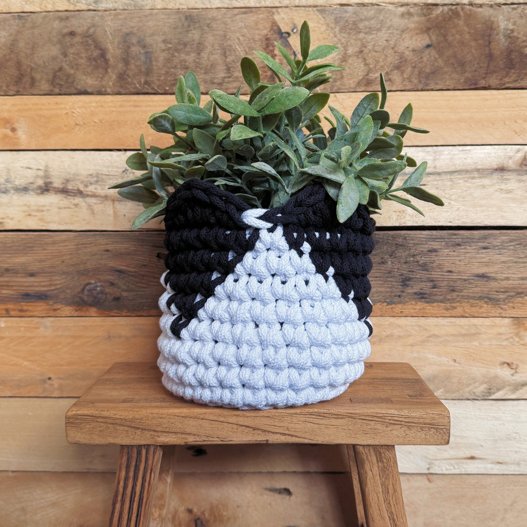A black and white crocheted planter basket holding a green leafy plant, displayed on a rustic wooden stool against a wooden plank wall.