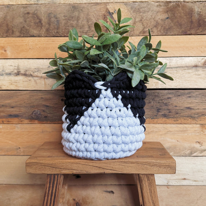 A black and white crocheted planter basket holding a green leafy plant, displayed on a rustic wooden stool against a wooden plank wall.